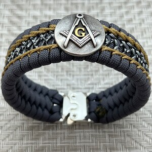 May include: A gray paracord bracelet with a gold and silver Masonic symbol charm and a silver clasp.