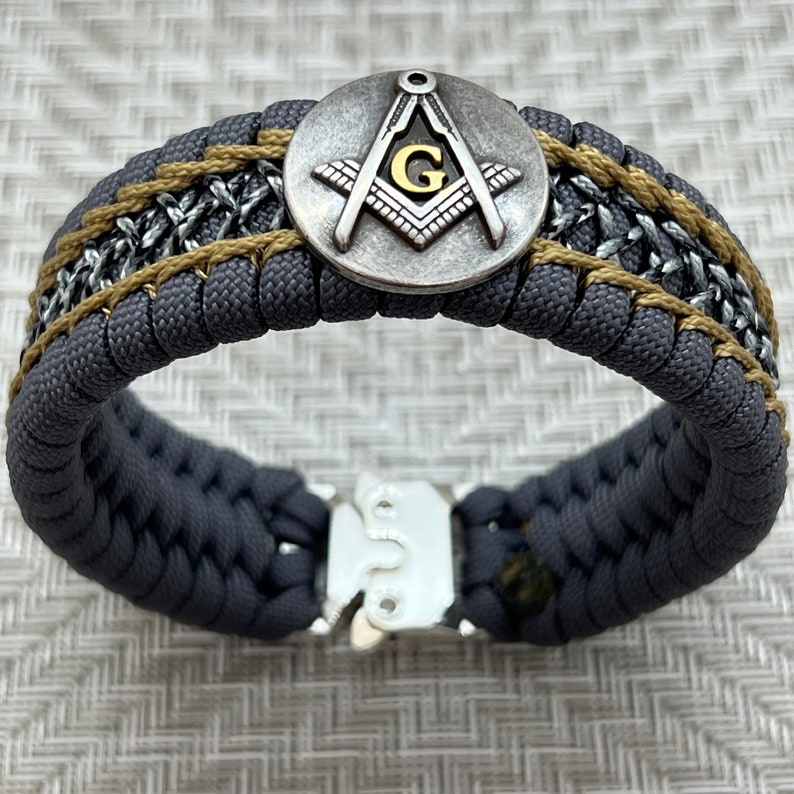 May include: A gray paracord bracelet with a gold and silver Masonic symbol charm and a silver clasp.