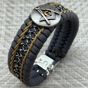 May include: A gray and gold paracord bracelet with a silver compass charm featuring a gold letter "G" in the center. The bracelet has a silver clasp.
