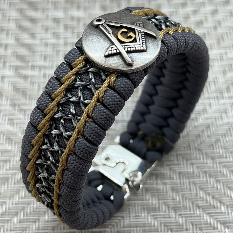 May include: A gray and gold paracord bracelet with a silver compass charm featuring a gold letter "G" in the center. The bracelet has a silver clasp.