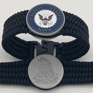 Op de afbeelding: Twee zwarte paracordarmbanden. Eén armband heeft een cirkelvormig embleem met de tekst "United States Navy" en een marineblauwe rand. De andere armband heeft een cirkelvormig embleem met een slang en de tekst "Don't Tread on Me".