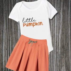 May include: A white bodysuit with the text "little pumpkin."  A coral-colored skirt with a drawstring waist and a green drawstring.