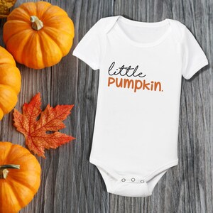 May include: White baby onesie with short sleeves and the text "little pumpkin." in black and orange lettering. The onesie is lying on a wooden surface with fall-colored leaves and pumpkins.