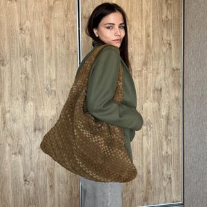 Puede incluir: Un bolso hobo grande y holgado en un tono marrón intenso, con un patrón de superficie tejido y texturizado. El bolso tiene una correa ancha para el hombro y una silueta suave y sin estructura que cuelga naturalmente del cuerpo.