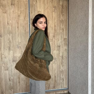 May include: A woman wearing a green blazer and gray pants is holding a large brown woven tote bag.