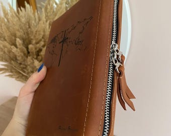 Puede incluir: Una funda de Biblia de cuero marrón con un mapa del mundo y un diseño de cruz. La funda tiene un cierre de cremallera plateado y una borla. El texto "Faith Forever" está impreso en la parte inferior.