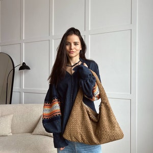 May include: A large, woven brown shoulder bag with a soft, slouchy design. The bag has a wide strap and is carried over the shoulder. The bag is made of a woven material and has a casual, everyday style.