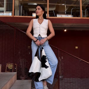 May include: A white sleeveless button-down vest, light blue denim jeans, and beige loafers paired with a large hobo-style shoulder bag featuring a black and white cow print pattern. The bag has a slouchy silhouette and a wide strap for carrying.