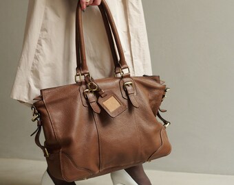 May include: A large brown pebbled leather tote bag with dual top handles, brass-toned metal hardware, buckle strap details on the front, and a small matching leather luggage tag hanging from one of the handle rings.