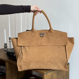May include: A large tan suede tote bag featuring two top handles, a wide fold-over flap, and a silver-toned metal hardware clasp centered on the front. The bag has a soft, textured surface and a simple, structured silhouette with side gussets.