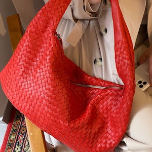 May include: A large, bright red woven leather shoulder bag with a zipper closure. The bag has a wide strap and a soft, slouchy shape. The bag is held by a person wearing a beige trench coat.