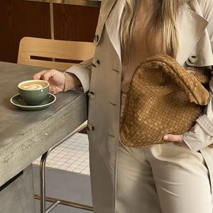 Può includere: Una donna con un trench beige e pantaloni beige tiene una borsa intrecciata di colore marrone. È seduta a un tavolo con una tazza di caffè.