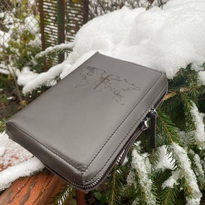 May include: A gray leather zippered case with a world map and a cross embossed on the front. The case is sitting on a wooden railing covered in snow.