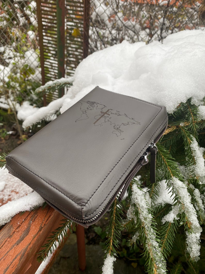 May include: A gray leather zippered case with a world map and a cross embossed on the front. The case is sitting on a wooden railing covered in snow.