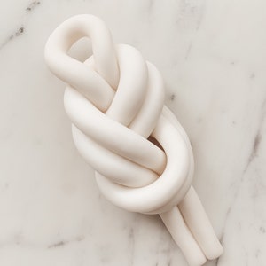 May include: A white ceramic knot sculpture, resembling a rope tied in a complex knot. The sculpture is smooth and has a matte finish.