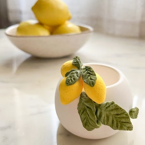 May include: A white ceramic planter with a round shape and a small cluster of yellow lemons and green leaves. The lemons and leaves are made of polymer clay.