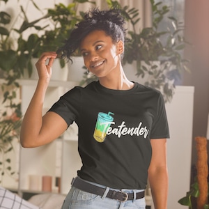 May include: Black t-shirt with a colorful graphic of a beverage and the word "Teatender" in white script. The shirt is short-sleeved and has a crew neck. The model is wearing a black belt and blue jeans.