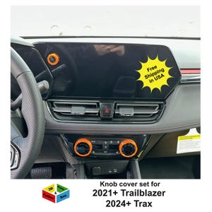 May include: Orange knob covers installed on a car dashboard. The covers are on the climate control knobs and the dashboard's start/stop button. The image includes the text "Free Shipping in USA" and "Knob cover set for 2021+ Trailblazer 2024+ Trax."