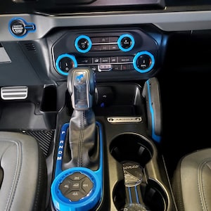 May include: Interior view of a car's dashboard and console. The dashboard features a black and gray design with blue accents around the controls. The gear shift and surrounding console are also accented with blue. A key fob with the word "Bronco" is in the cup holder.