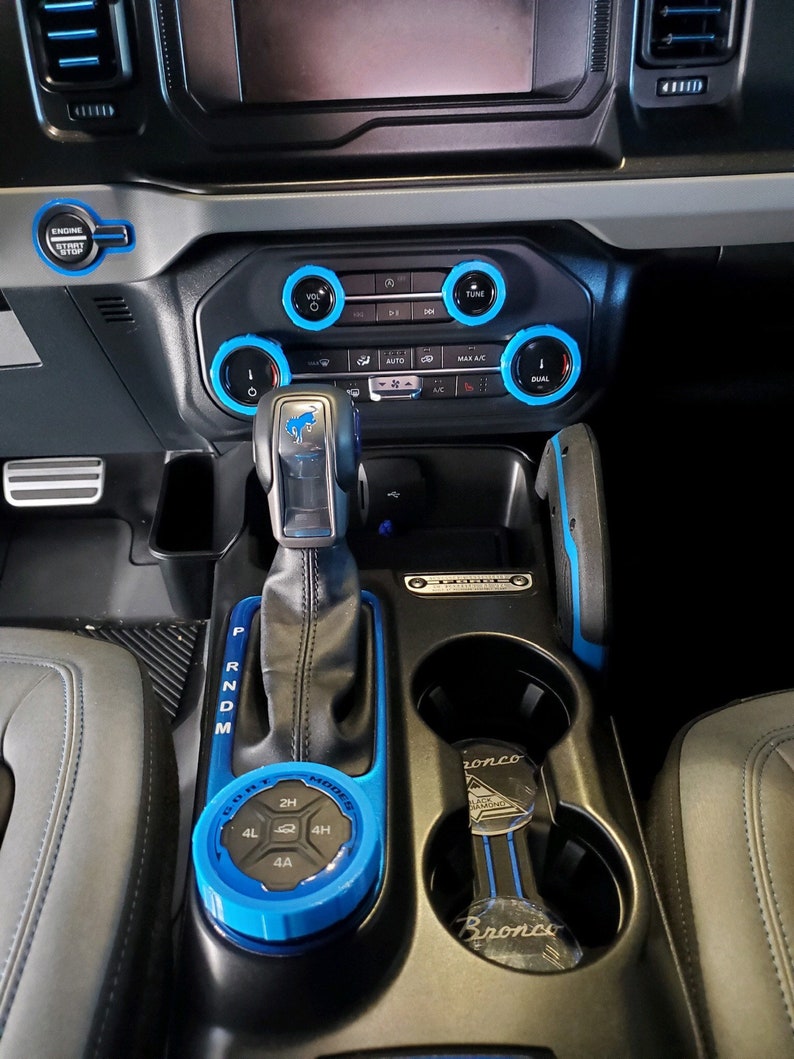May include: Interior view of a car's dashboard and console. The dashboard features a black and gray design with blue accents around the controls. The gear shift and surrounding console are also accented with blue. A key fob with the word "Bronco" is in the cup holder.