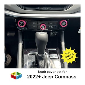 May include: A set of pink knob covers installed on a car's dashboard. The covers are designed for a 2022+ Jeep Compass. The image also shows the car's gear shift, air conditioning controls, and the text "Free Shipping in USA".