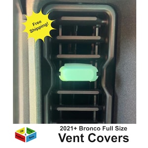 May include: A light green rectangular vent cover installed on a black vehicle air conditioning vent. The cover is positioned horizontally over the center slat of the vent. The text 2021+ Bronco Full Size Vent Covers is visible below the vent.