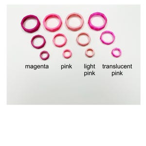 May include: An assortment of pink rings in varying sizes and shades, including magenta, pink, light pink, and translucent pink. The rings are arranged on a white background, with the color names labeled below each group.