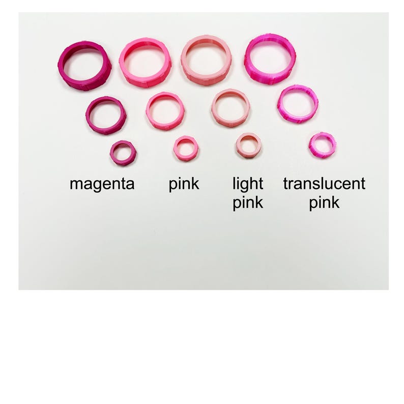 May include: An assortment of pink rings in varying sizes and shades, including magenta, pink, light pink, and translucent pink. The rings are arranged on a white background, with the color names labeled below each group.