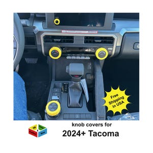 May include: Interior view of a car dashboard with yellow knob covers. The dashboard is black and gray, with a gear shift and air vents. Text on the image reads "knob covers for 2024+ Tacoma".