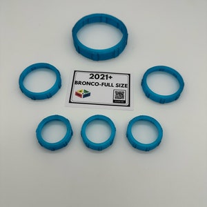 May include: Several turquoise-colored, circular objects with a ridged design are arranged on a white surface. A rectangular card with the text "2021+ BRONCO-FULL SIZE" is also present, along with a QR code.