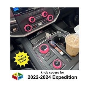 May include: Interior car detail featuring bright pink knob covers for a 2022-2024 Expedition. The image shows the car's dashboard and center console with the custom covers installed. A coffee drink and a pen are also visible.