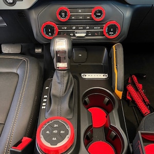 May include: Interior car accessories featuring red accents. The image shows a gear shift, cup holders, and control knobs with red trim. The dashboard includes buttons labeled "VOL", "TUNE", and "DUAL". The gear shift knob has the text "MODES" and "2H, 4L, 4H, 4A".