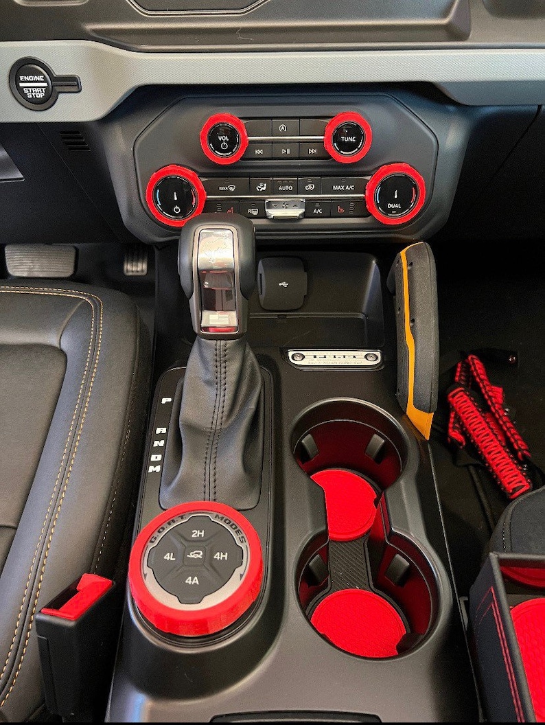 May include: Interior car accessories featuring red accents. The image shows a gear shift, cup holders, and control knobs with red trim. The dashboard includes buttons labeled "VOL", "TUNE", and "DUAL". The gear shift knob has the text "MODES" and "2H, 4L, 4H, 4A".