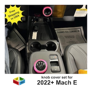 May include: Interior car accessories, including a pink knob cover set for a 2022+ Mach E. The image shows the car's console with the gear shift knob and other controls. The text "Free Shipping in USA" is visible.