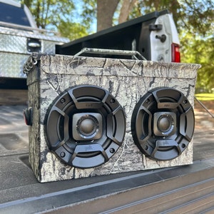 May include: A camouflage-patterned speaker box with two black Polk Audio speakers. The speakers have a black grill and a silver Polk Audio logo.