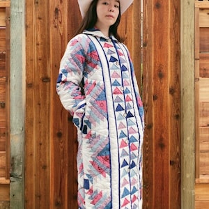 May include: A long, quilted coat with a geometric pattern in shades of white, pink, and blue. The coat has a button-up front and a collar. The person is wearing a white hat. The coat is made of a soft, quilted material.