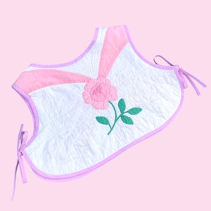 May include: A white quilted tote bag with a pink rose applique and green leaves. The bag has pink accents and ties, with a light pink border. The background is a solid light pink.
