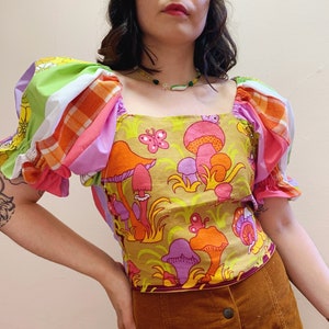 May include: A light brown top with a square neckline and puff sleeves. The top features a colourful mushroom and butterfly print in shades of pink, orange, purple, and green.