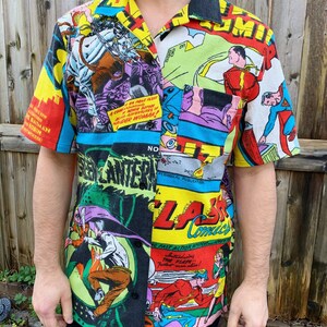 May include: A short-sleeved button-up shirt with a colorful comic book print featuring superheroes like Superman, Batman, Robin, Wonder Woman, Green Lantern, and The Flash. The shirt has a black collar and a pocket on the left side.