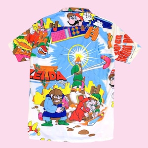 Può includere: Una camicia bianca con bottoni e una stampa colorata con personaggi dei videogiochi Super Mario Bros. e The Legend of Zelda. La stampa include Mario, Luigi, Link e altri personaggi dei giochi.