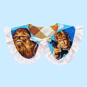 Peut inclure: Un col en dentelle blanche avec un insert en tissu bleu et marron représentant des images de Chewbacca et Han Solo de Star Wars. L'insert en tissu a un motif à carreaux en arrière-plan.