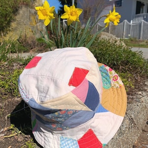 Op de afbeelding: Een witte bucket hat gemaakt van patchwork stof met verschillende kleuren en patronen, waaronder rood, blauw, geel, roze en groen. De hoed ligt op een rots.