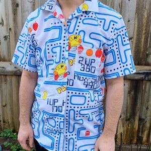 May include: A white short-sleeved button-up shirt with a blue and white Pac-Man pattern. The shirt features Pac-Man characters, ghosts, and the Pac-Man maze. The text "2 UP 380" and "1 UP 170" are printed on the shirt.