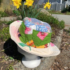 Puede incluir: Un sombrero de parches blanco y crema con estampado floral. El sombrero está hecho de diferentes telas y colores, incluyendo verde, rosa, azul y amarillo. El sombrero está sentado en una roca con plantas verdes en el fondo.