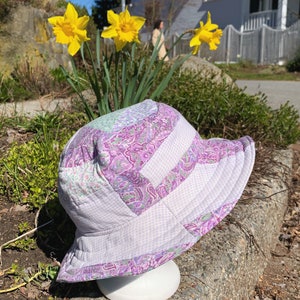 Puede incluir: Un sombrero de cubo de patchwork blanco y morado con un patrón de cuadros. El sombrero está sobre una superficie de piedra.