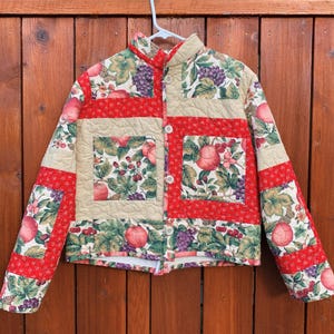 May include: A quilted jacket with a red and beige patchwork design. The fabric features a floral print with apples, grapes, and berries.