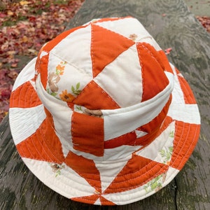 Puede incluir: Un sombrero de cubo de patchwork blanco y naranja con estampado floral. El sombrero está hecho de tela y tiene un ala ancha.