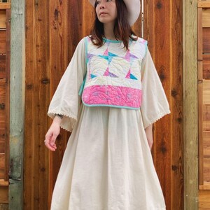 May include: A cream-coloured dress paired with a white quilted vest, adorned with pink, blue, and white star patterns. The dress features long sleeves with lace trim, complemented by a matching white hat. The backdrop is a wooden surface.