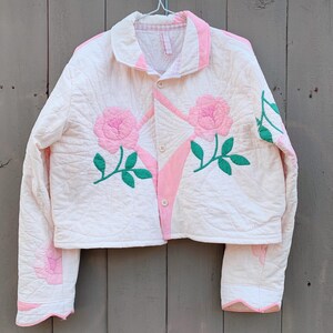 May include: A quilted jacket in cream with pink and green floral appliqués. The jacket features a collar, button closure, and long sleeves. Pink accents trim the collar, front, and sleeves. The floral design includes pink roses and green leaves.