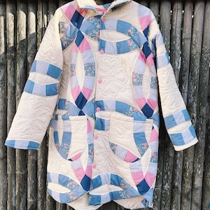 Pode incluir: Uma jaqueta acolchoada de cor creme com um design de patchwork rosa e azul. A jaqueta tem um fecho de botões e uma gola.